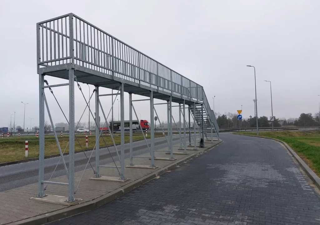 Metalowa kładka dla pieszych ze schodami, ustawiona nad chodnikiem, prowadzi równolegle do ulicy obok ronda; w tle widoczna droga, auta ciężarowe i znaki drogowe, otoczenie miejskie z szarą pogodą.