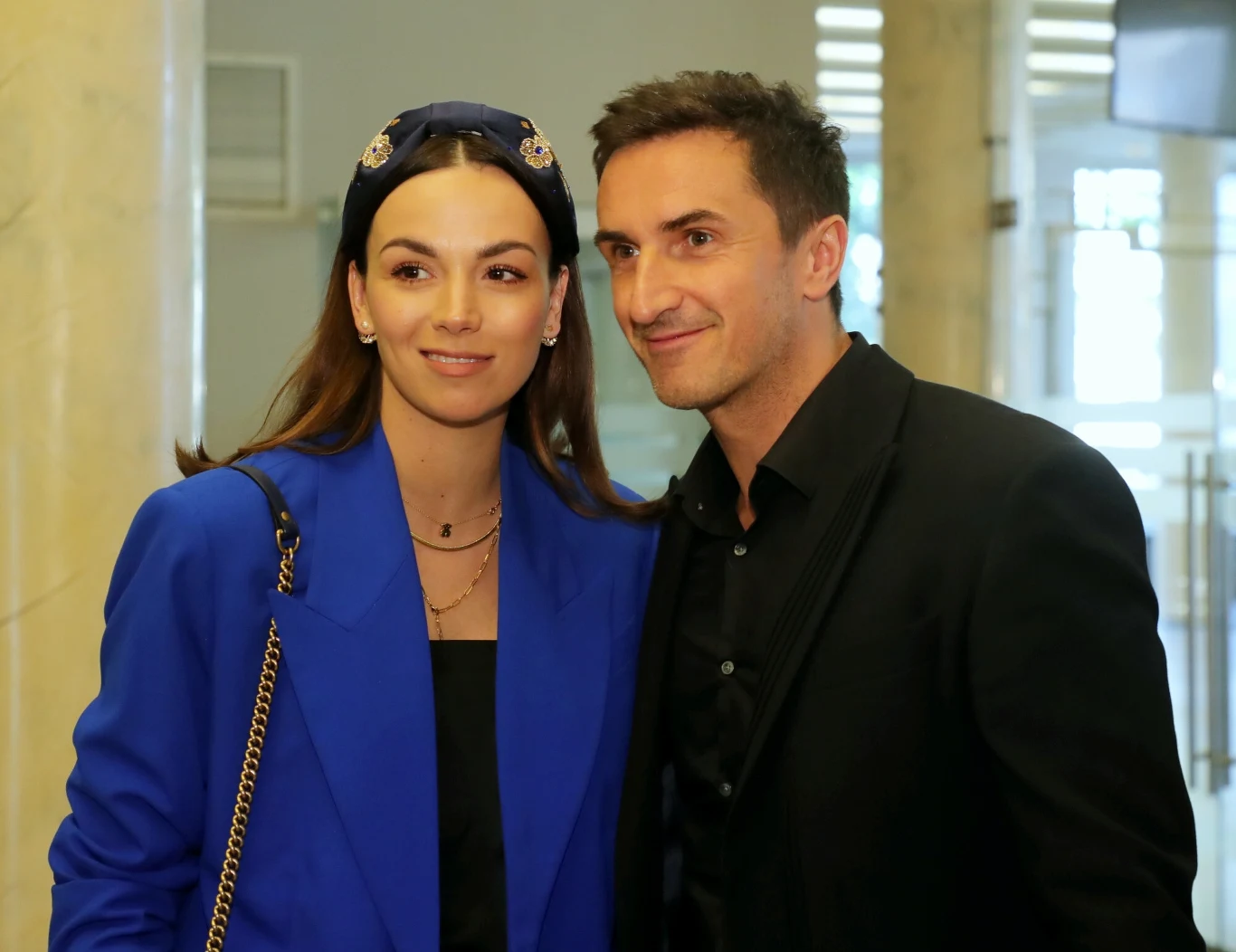 Paulina Krupińska-Karpiel i jej mąż Sebastian Karpiel-Bułecka Młoda kobieta w niebieskiej marynarce z opaską we włosach oraz mężczyzna w czarnej marynarce stoją razem, uśmiechając się do zdjęcia w jasnym wnętrzu z wysokimi kolumnami