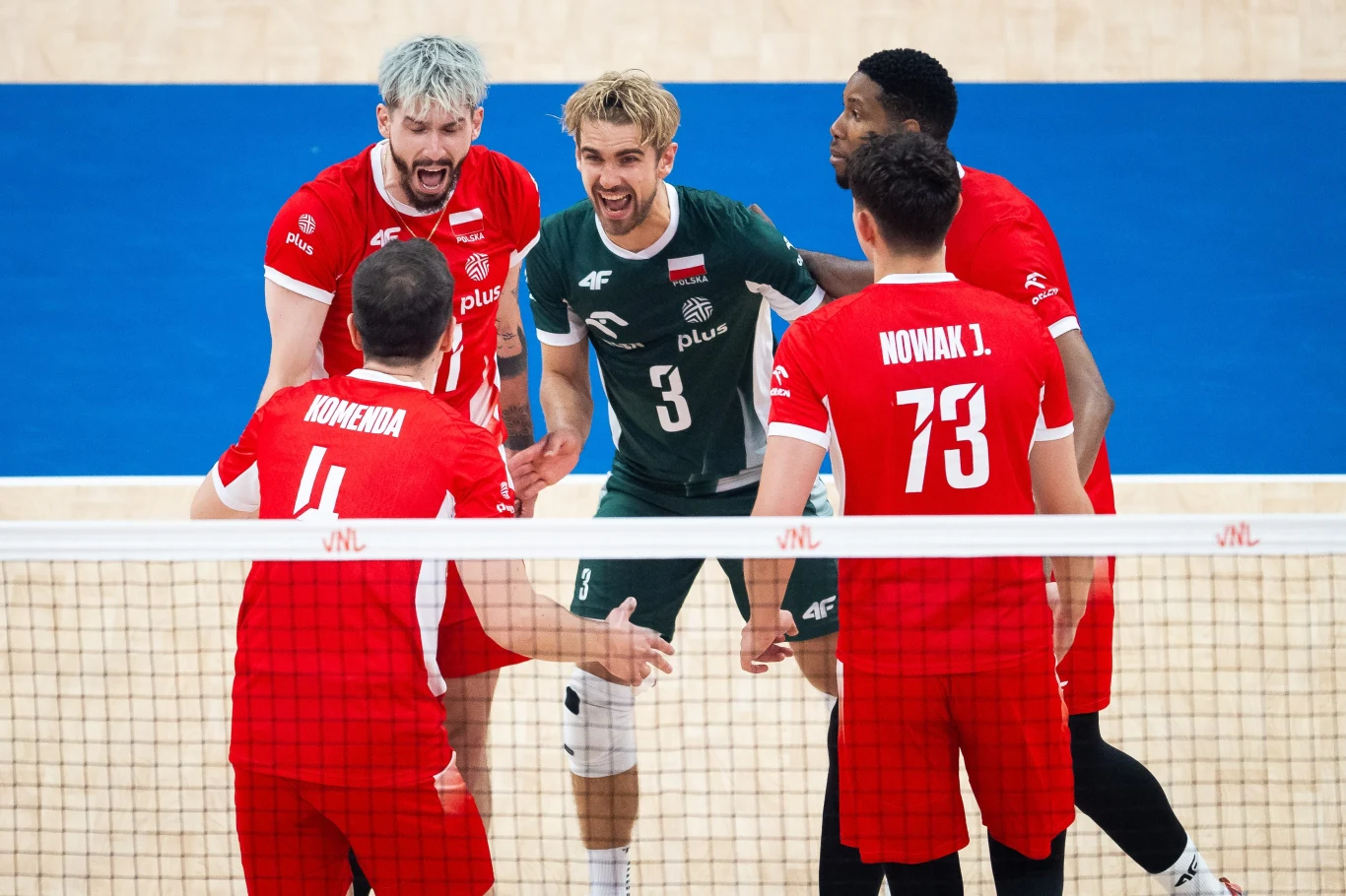 Reprezentacja Polski siatkarzy w meczu Ligi Narodów Pięciu siatkarzy w sportowych strojach cieszy się wspólnie po zdobyciu punktu podczas meczu siatkówki na hali sportowej, na pierwszym planie widoczna jest siatka, a tło stanowi niebieska wykładzina boiska.
