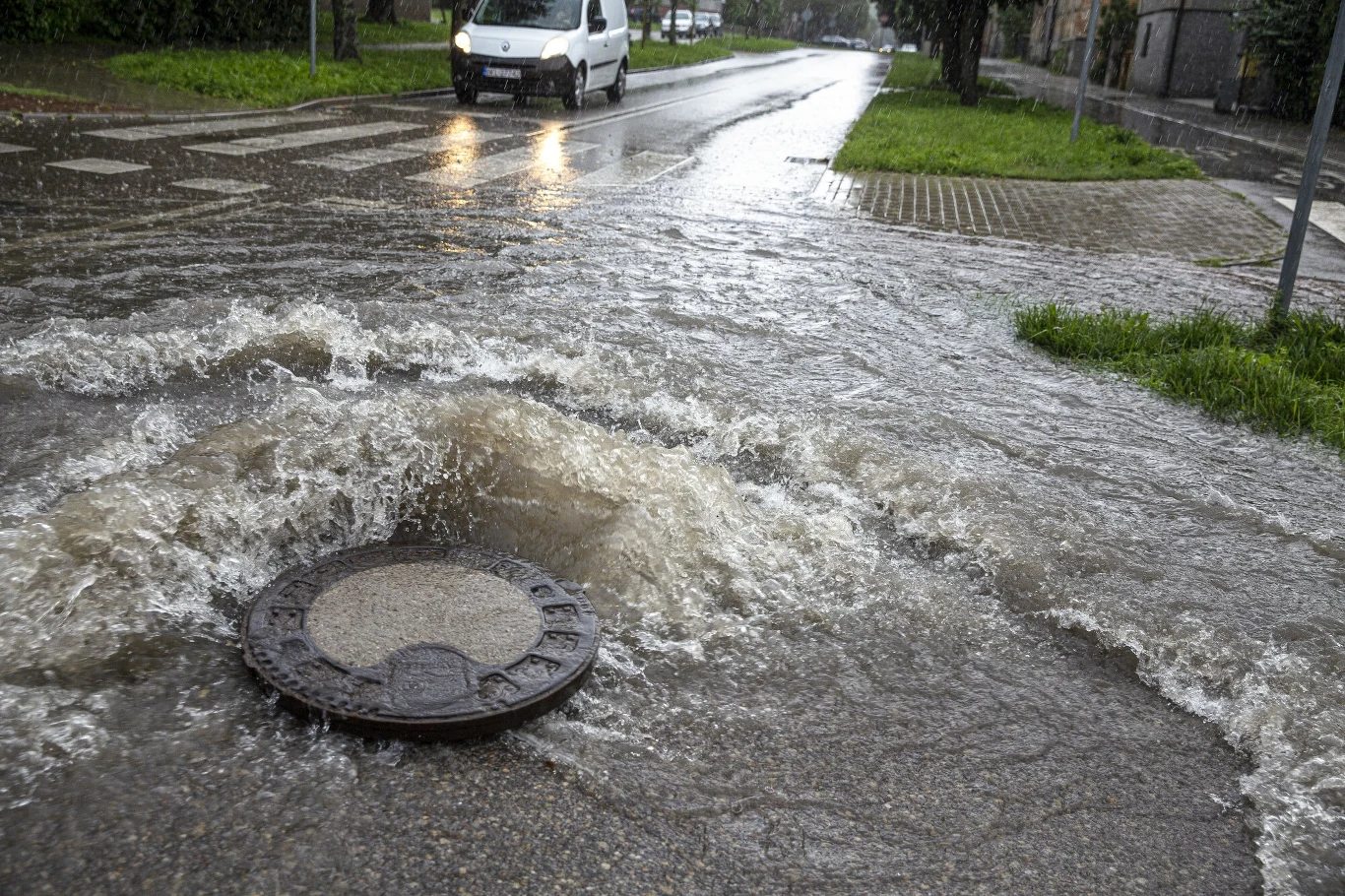 Woda wypływająca z kanalizacji przez studzienkę zalewa ulicę, a w tle widoczny jest samochód jadący przez zalany odcinek drogi.