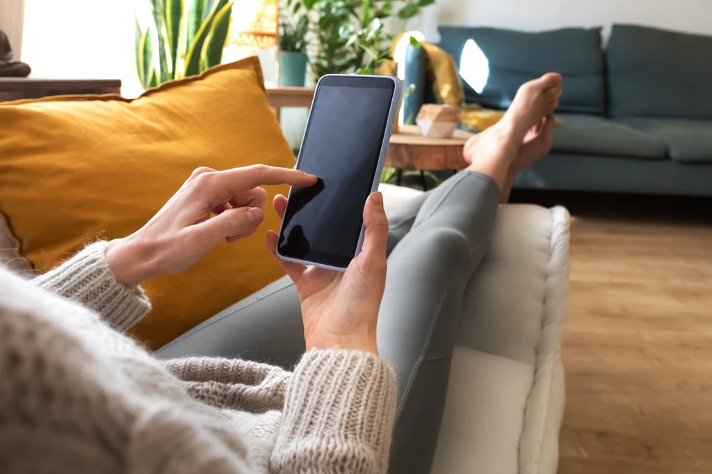 Każdy z nas może zrobić zakupy w Biedronce-widmo, jednak nie osobiście Osoba siedzi wygodnie na kanapie z żółtą poduszką, przeglądając smartfon. W tle widać zielone rośliny doniczkowe i nowoczesny wystrój salonu, przeważają ciepłe kolory i przytulna atmosfera.