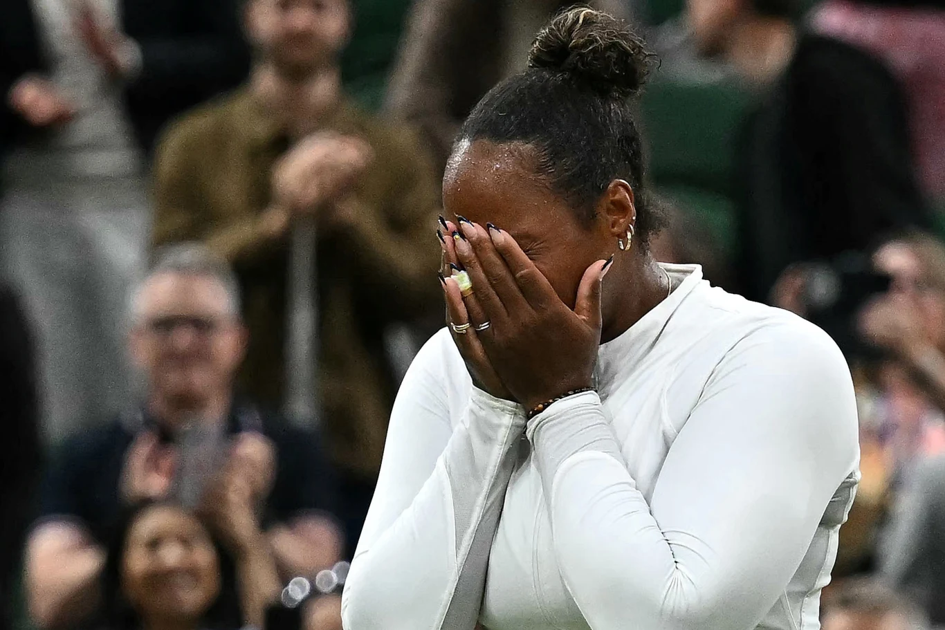 Kobieta ubrana na biało zasłania twarz dłońmi w geście silnych emocji, na drugim planie rozmazana publiczność bijąca brawo.