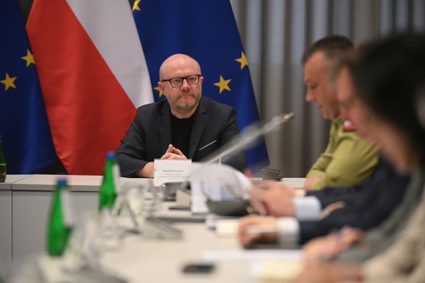 Maciej Duszczyk Urzędnik siedzący przy stole konferencyjnym, przed nim tabliczka imienna, w tle flagi Polski oraz Unii Europejskiej, po lewej kilka butelek wody, po prawej grupa osób skupionych na dokumentach.