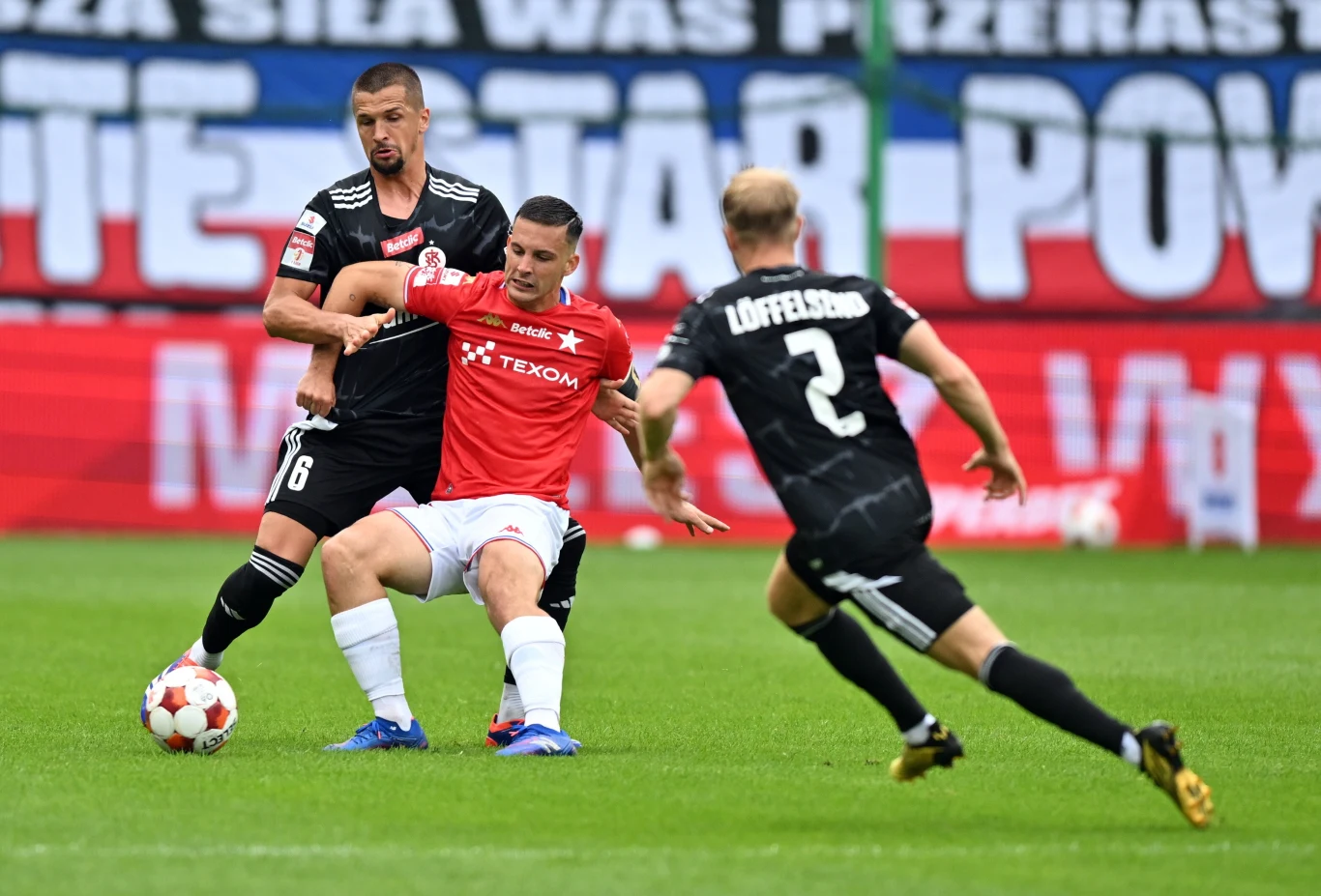 Mecz Wisła Kraków - ŁKS Łódź Trzech piłkarzy walczy o piłkę na zielonej murawie, dwóch ubranych w czarne stroje i jeden w czerwonej koszulce, w tle kolorowy transparent kibiców.
