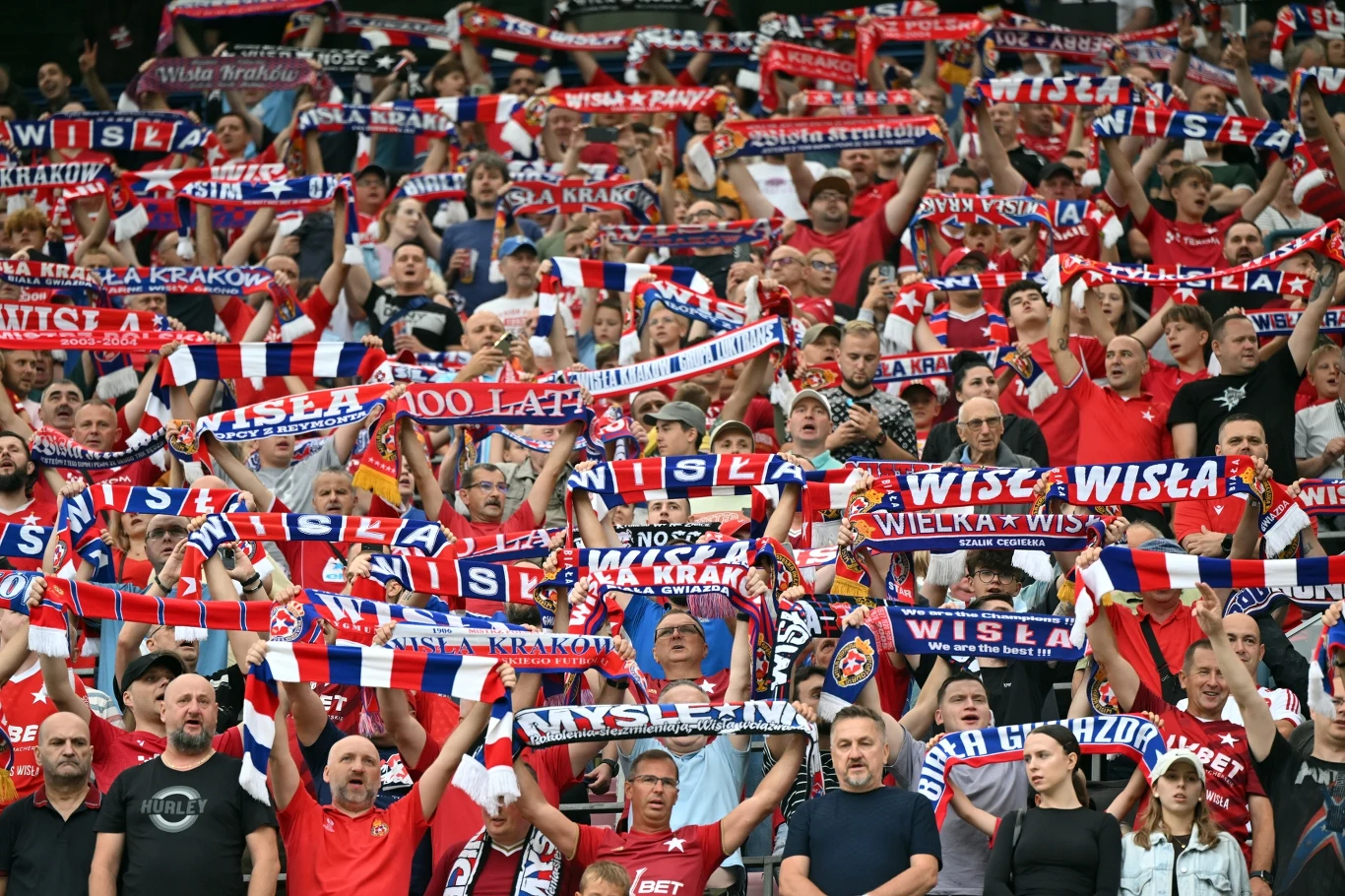 Kibice Wisły Kraków Tłum kibiców ubranych w czerwono-niebiesko-białe barwy, trzymających szaliki z napisami wspierającymi klub Wisła Kraków, wyrażających entuzjazm i sportową solidarność.