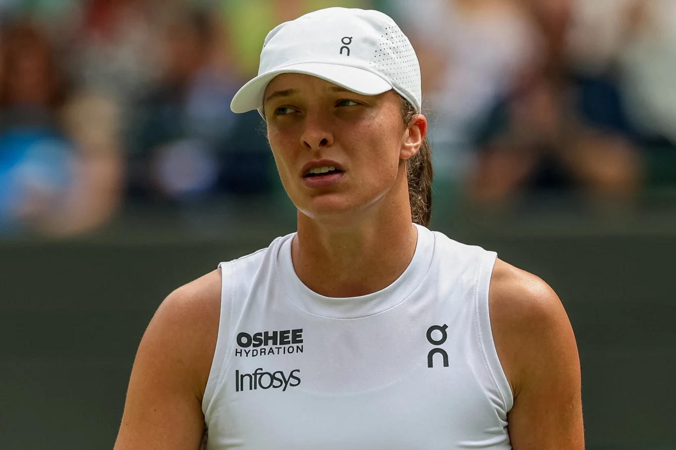 Tenisistka ubrana w biały sportowy strój i czapkę z daszkiem stoi na korcie tenisowym, widoczne są logotypy sponsorów na jej koszulce; tło jest rozmyte i sugeruje obecność widowni.