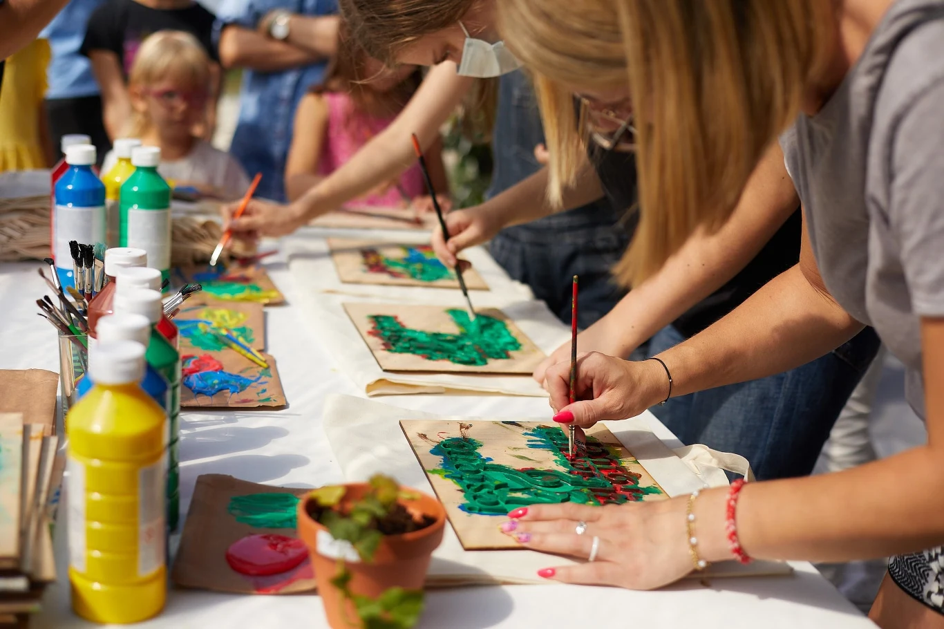 Grupa osób maluje obrazy na kartkach papieru rozłożonych na stole, korzystając z kolorowych farb i pędzli, wokół widoczne butelki z farbami oraz pędzle, w tle inne osoby obserwują zajęcia.