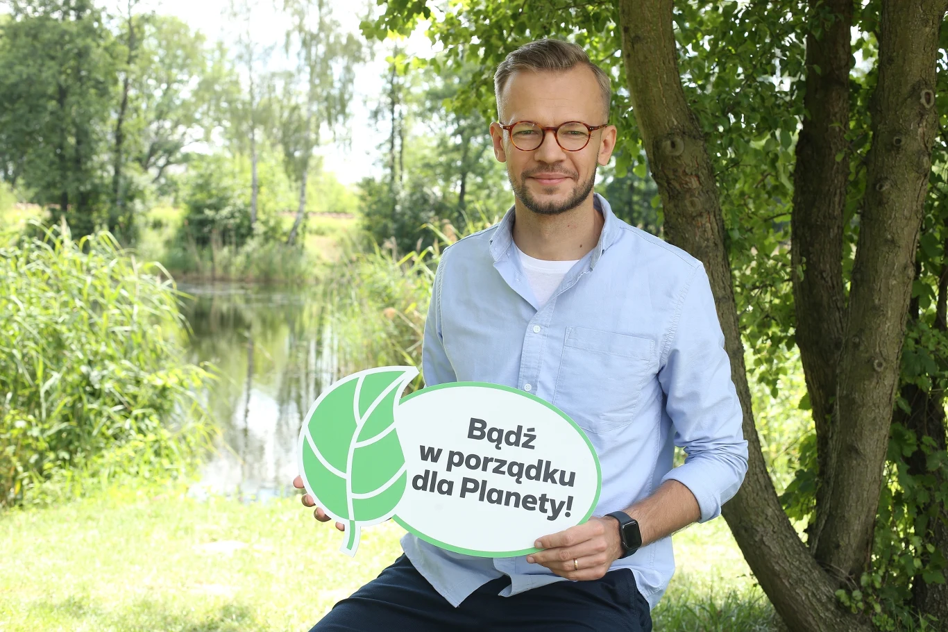 Mężczyzna w okularach siedzi na ławeczce przy jeziorze, trzymając tabliczkę z napisem „Bądź w porządku dla Planety!”, wokół bujna, zielona roślinność.