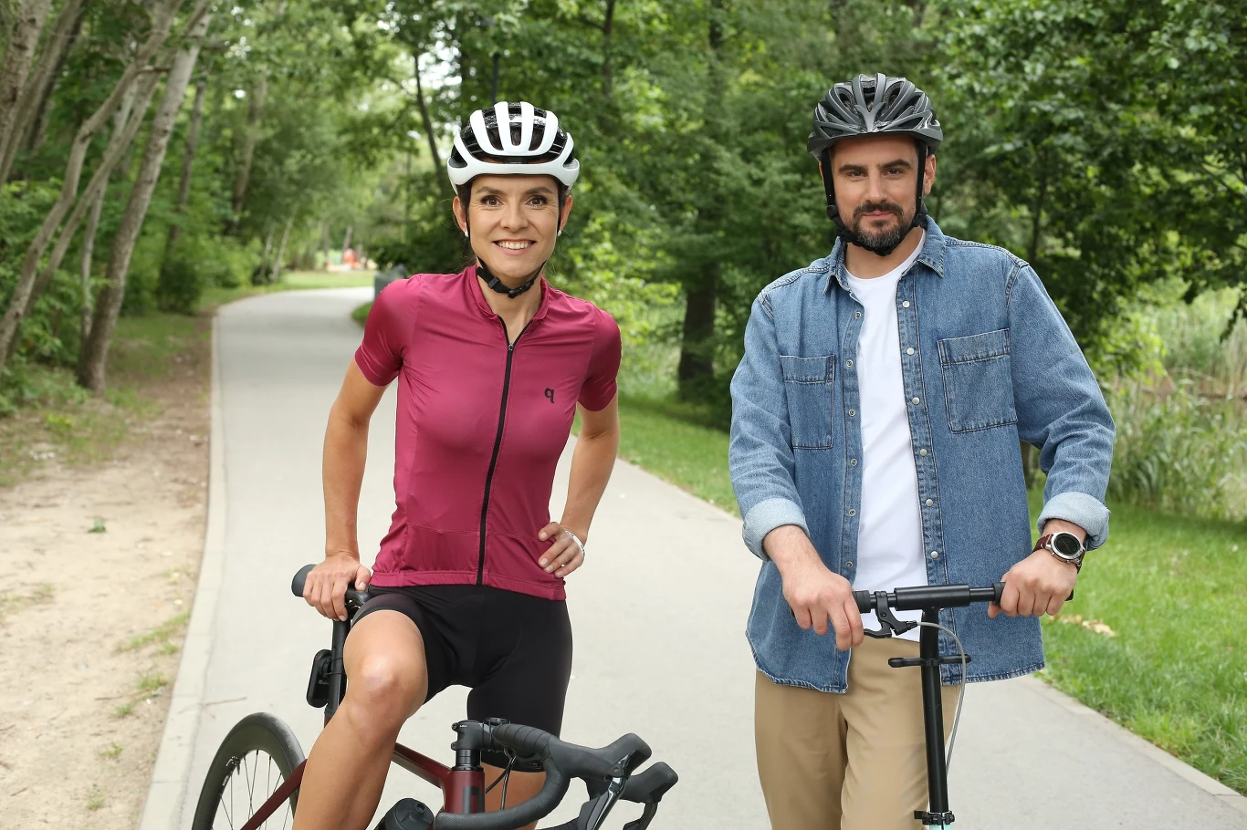 Dwie osoby w kaskach rowerowych stoją na ścieżce rowerowej w lesie, jedna z nich ubrana w sportowy strój i trzyma rower, druga w casualowej koszuli i spodniach również opiera się o rower, w tle zielone drzewa.
