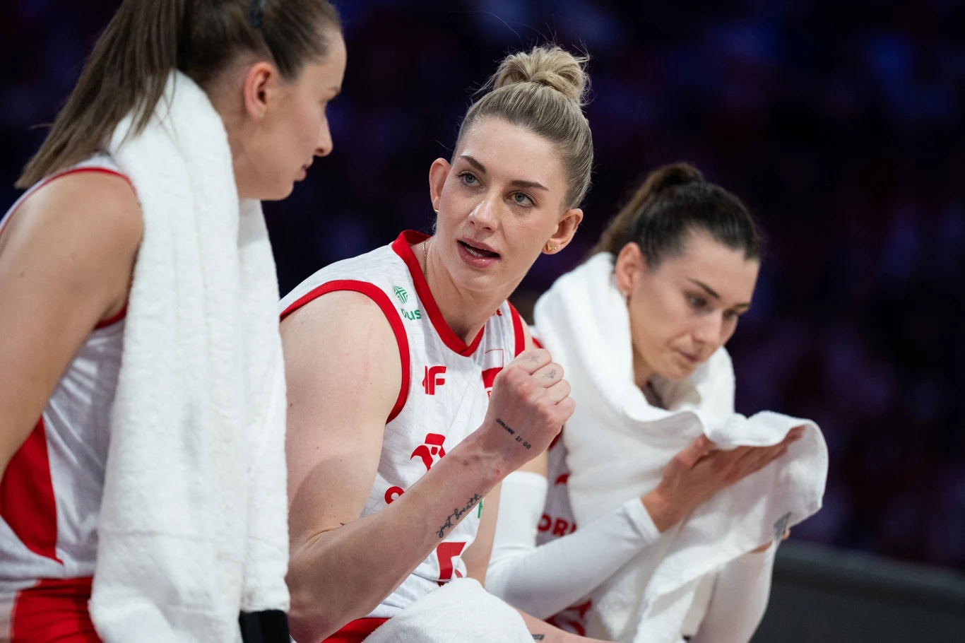 Magdalena Stysiak (w środku). Po lewej Magdalena Jurczyk, a po prawej Paulina Damaske Trzy koszykarki w białych strojach z czerwonymi akcentami i biało-czerwonymi ręcznikami siedzą na ławce podczas meczu, prowadząc rozmowę i odpoczywając po grze