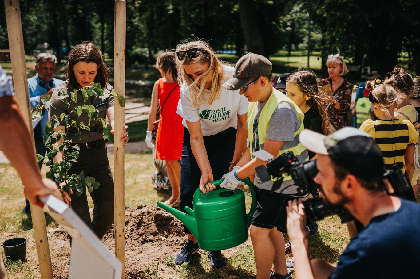 Dzieci i dorośli wspólnie sadzą drzewo w parku, podlewając młodą roślinę dużą zieloną konewką, otoczeni są przez innych uczestników wydarzenia i operatora kamery dokumentującego akcję.