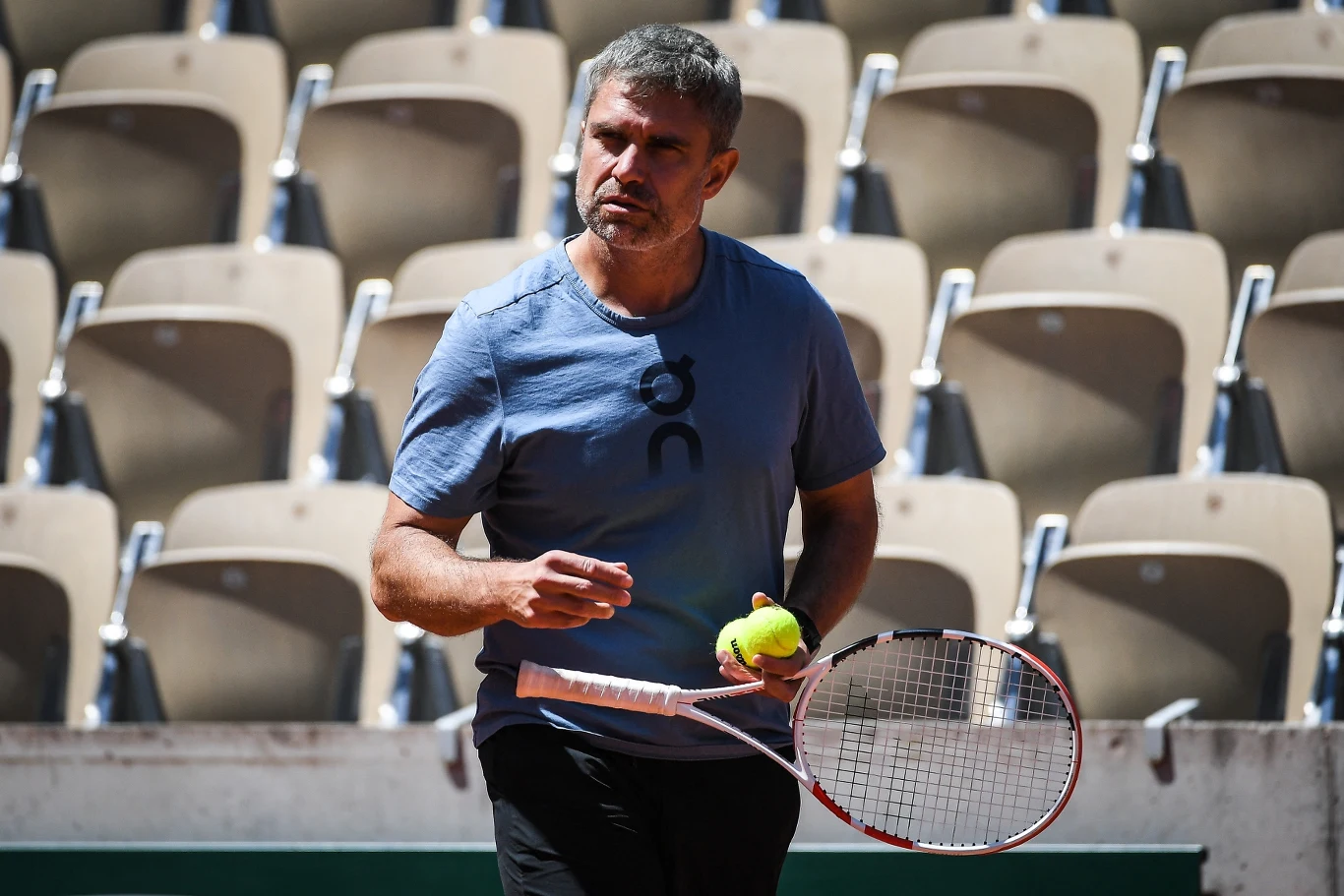 Tomasz Wiktorowski Mężczyzna w sportowej koszulce trzymający rakietę tenisową i dwie piłki tenisowe, na tle pustych trybun kortu tenisowego.