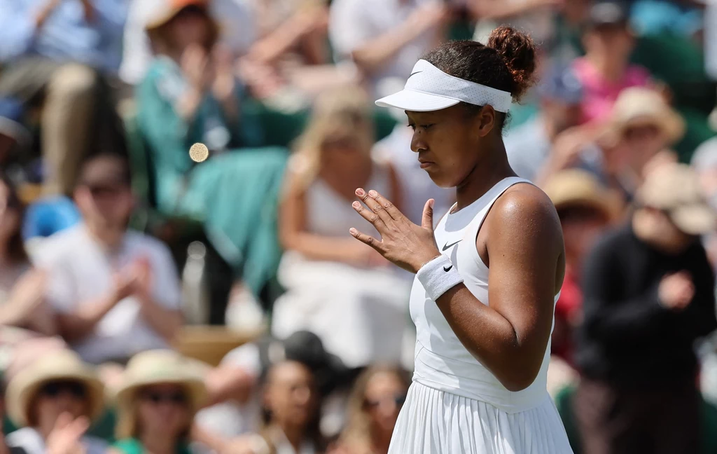 Naomi Osaka Tenisistka ubrana w biały strój sportowy i daszek stoi na korcie, koncentrując się przed serwisem, w tle rozmyta publiczność na trybunach.