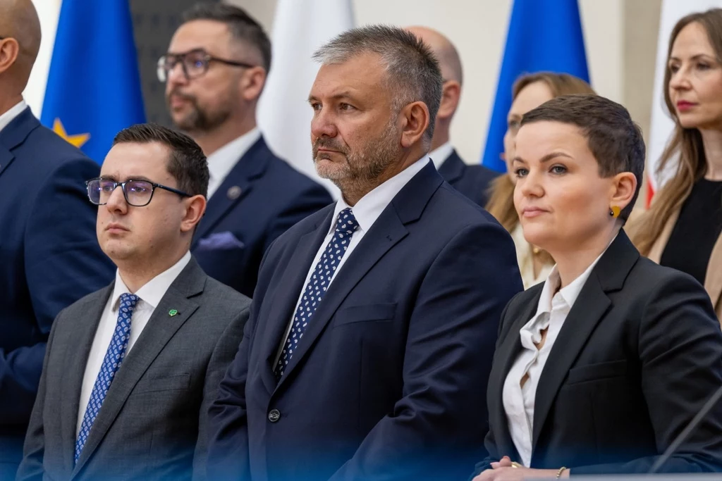 Waldemar Żurek ma przede wszystkim odzyskać kontrolę nad prokuraturą Grupa poważnie wyglądających osób w garniturach i formalnym stroju stoi w oficjalnym otoczeniu, w tle widoczne flagi państwowe oraz flaga Unii Europejskiej, sugerujące udział w ważnym wydarzeniu politycznym lub konferencji.