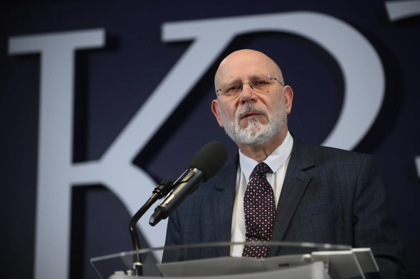 Maciej Świrski odwołany ze stanowiska przewodniczącego KRRiT Starszy mężczyzna z brodą i okularami w garniturze stoi przy mównicy, przemawiając do mikrofonu na tle dużych liter jako elementu wystroju sali.