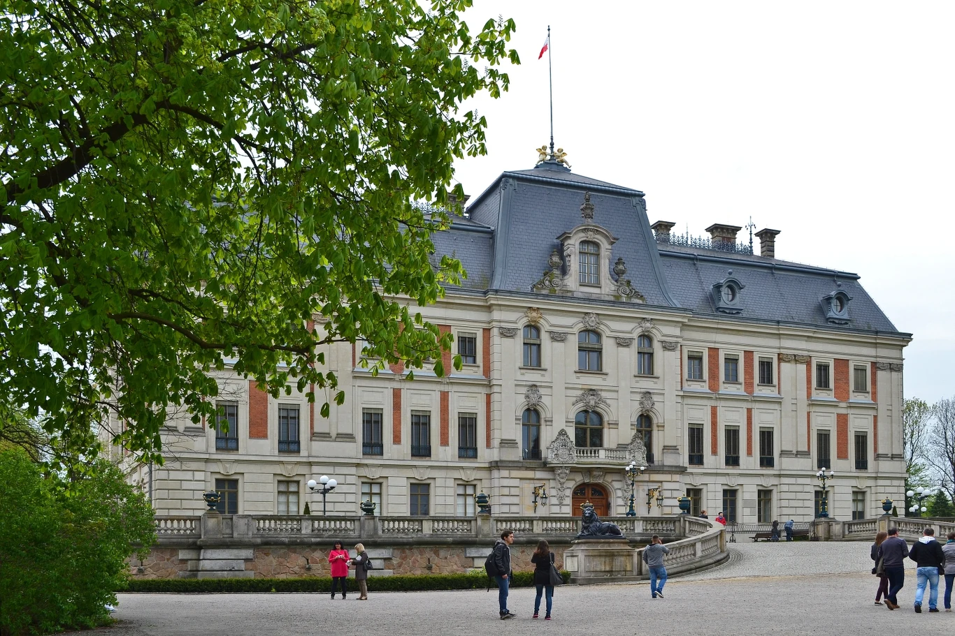 Okazały pałac w stylu barokowym, otoczony szerokim dziedzińcem, na którym znajdują się nieliczne osoby zwiedzające i spacerujące. W tle widoczne rozłożyste drzewo z zielonymi liśćmi, nadające zdjęciu wiosennego charakteru.
