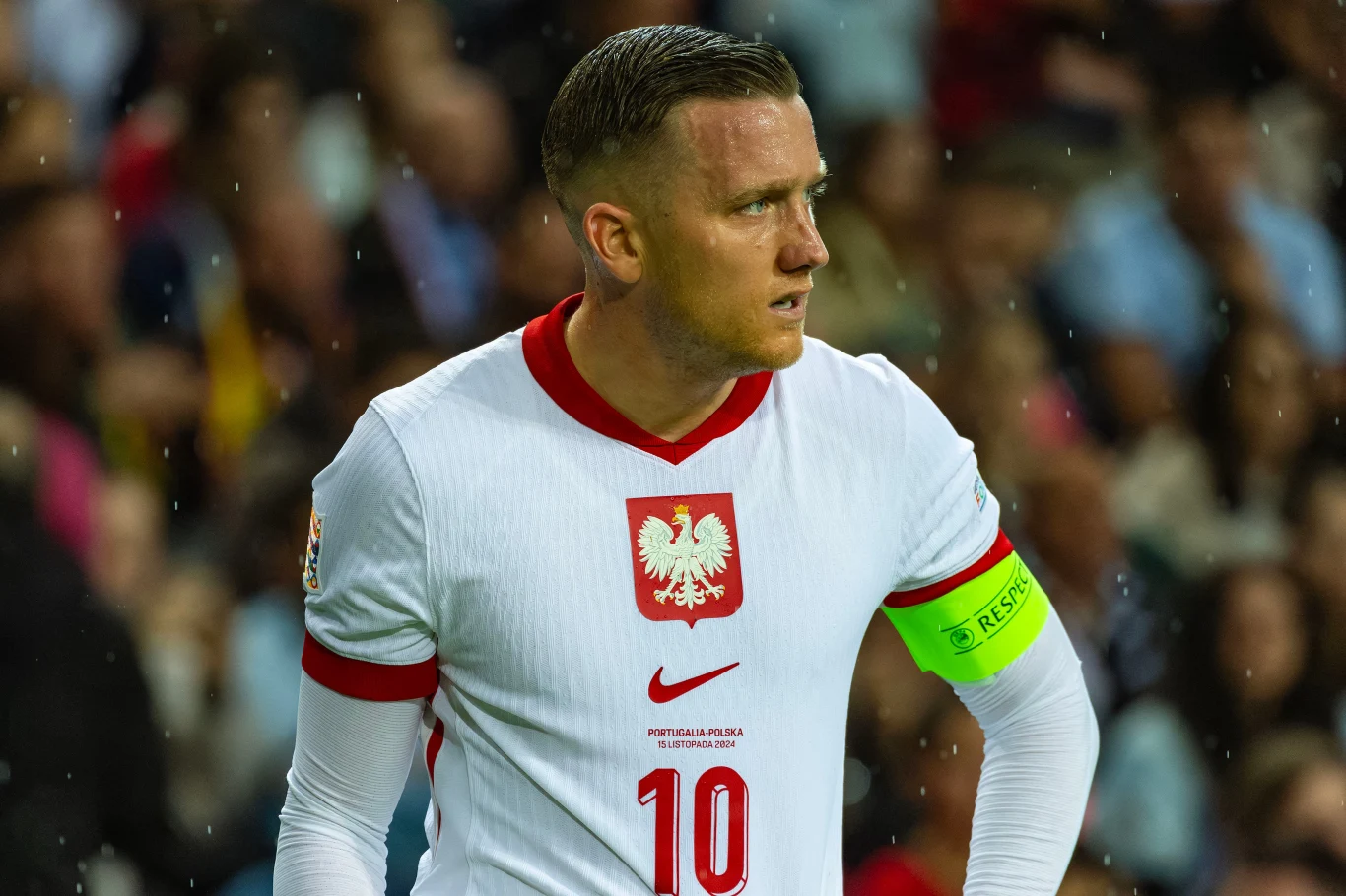 Piotr Zieliński Piłkarz w białej koszulce z numerem 10 i opaską kapitańską na ramieniu, podczas meczu, tłum kibiców w tle.