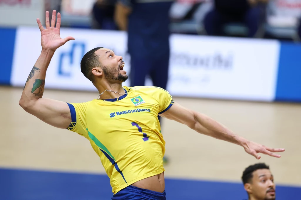 Zawodnik brazylijskiej reprezentacji siatkarskiej w żółto-zielonej koszulce wykonuje dynamiczny wyskok, przygotowując się do zagrania piłki podczas meczu, w tle widoczny jest fragment hali sportowej i inny zawodnik.