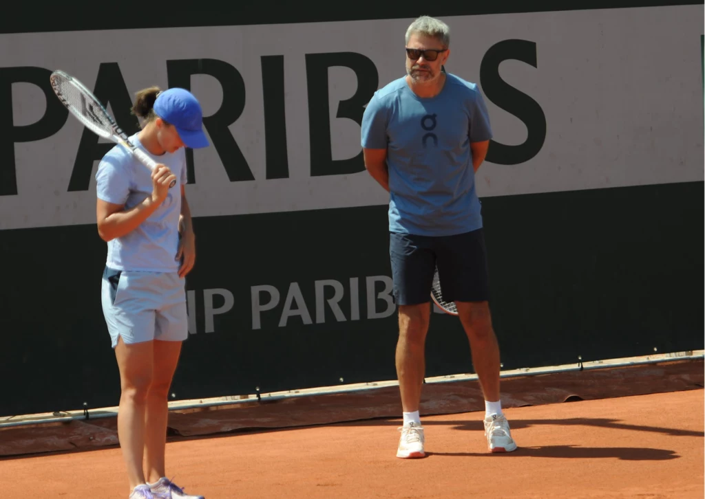 Iga Świątek i Tomasz Wiktorowski Kobieta w sportowym stroju i czapce z daszkiem trzymająca rakietę tenisową na korcie ziemnym, obok niej mężczyzna w okularach przeciwsłonecznych i niebieskiej koszulce, prawdopodobnie trener, oboje skupieni na rozmowie podczas treningu.