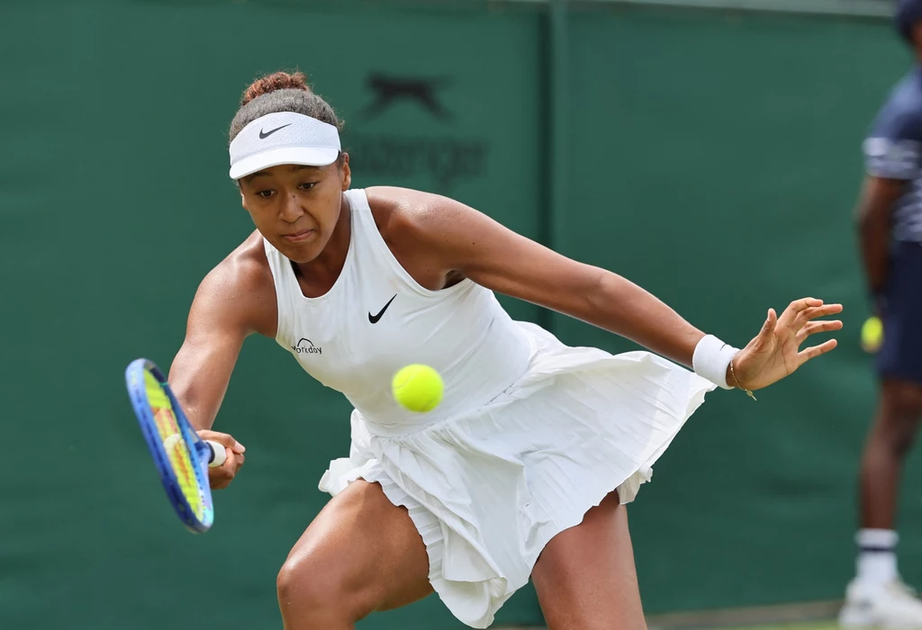 Naomi Osaka Tenisistka ubrana w białą sportową sukienkę dynamicznie odbija piłkę tenisową rakietą podczas meczu na zielonym korcie.