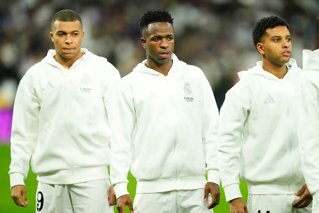 Kylian Mbappe, Vinicius Junior i Rodrygo Trzech piłkarzy ubranych w białe bluzy sportowe z herbem klubu, stojących obok siebie na murawie, skoncentrowanych przed rozpoczęciem meczu.