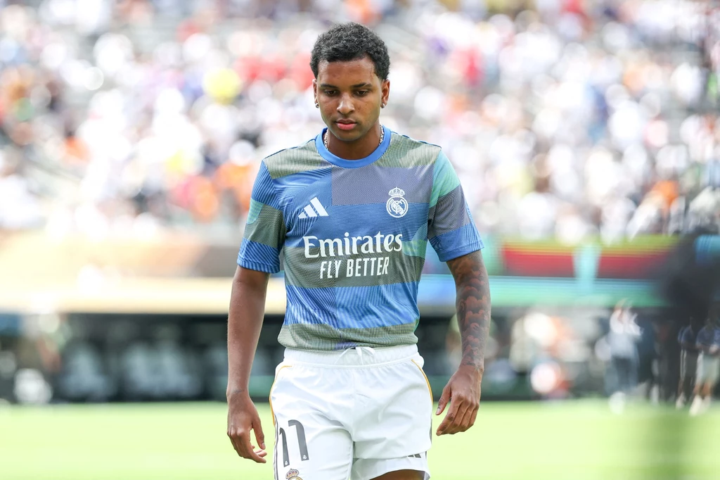 Rodrygo Młody piłkarz w niebiesko-zielonej koszulce treningowej Realu Madryt, z numerem 11 na spodenkach, stoi na murawie stadionu w trakcie treningu lub rozgrzewki, w tle rozmyte trybuny pełne kibiców.