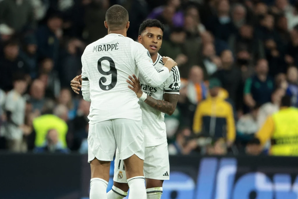 Kylian Mbappe i Rodrygo Dwóch piłkarzy w białych strojach obejmuje się na boisku podczas meczu piłkarskiego, w tle widoczni kibice na trybunach.