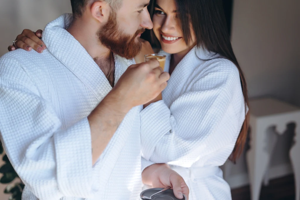 "Princess treatment" w związku Para ubrana w białe, hotelowe szlafroki, stojąca blisko siebie i uśmiechająca się, mężczyzna trzyma filiżankę z napojem, a kobieta obejmuje partnera.