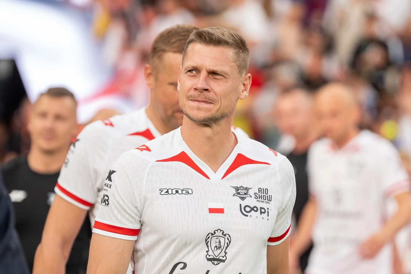 Łukasz Piszczek Mężczyzna w sportowej koszulce z logo i numerem, stojący na tle innych osób ubranych podobnie, atmosfera sugeruje udział w wydarzeniu sportowym, tłum w tle dodaje energii scenie.