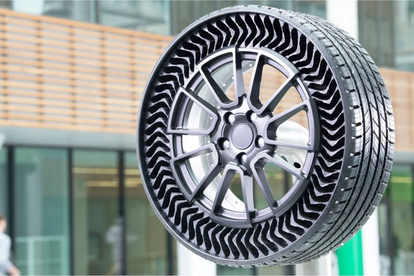 Michelin Uptis jest odporny na przebicie. Nowoczesna opona bezpowietrzna z widoczną innowacyjną strukturą boczną umożliwiającą elastyczność, osadzona na stylowej feldze aluminiowej, w tle szklane ściany budynku i elementy architektury miejskiej.