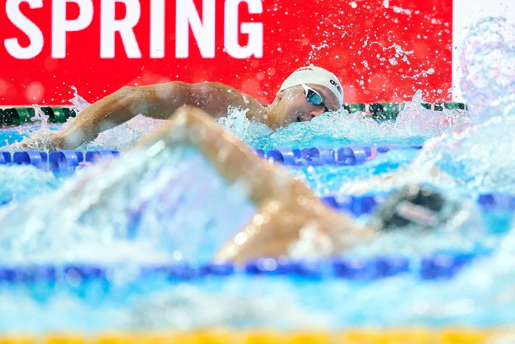 Krzysztof Chmielewski Dwóch pływaków rywalizujących w wyścigu na basenie, dynamiczne pluski wody, wyraźny napis SPRING w tle, intensywna atmosfera zawodów sportowych.