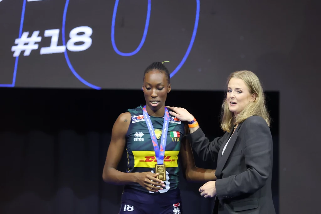 Liga Narodów siatkarek. Paola Egonu została wybrana do drużyny marzeń turnieju. Złoty medal wręczyła jej Joanna Wołosz Siatkarka w stroju reprezentacji Włoch odbiera medal od kobiety w eleganckim ubraniu, tło stanowi duży ekran z numerem 18.