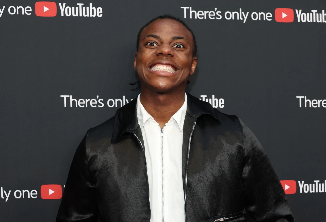 Młody mężczyzna z szerokim uśmiechem pozuje na tle ścianki z logotypami YouTube oraz napisem 'There's only one', ubrany w czarną satynową kurtkę i białą koszulę.