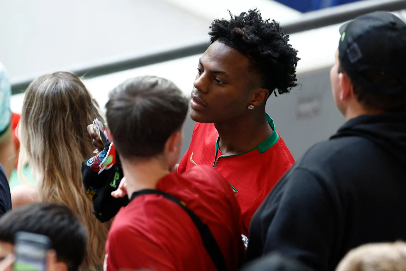 Młody mężczyzna w czerwonej koszulce sportowej z zielonymi elementami stoi wśród grupy osób, skupiony na czymś poza kadrem, wokół niego widać kilka innych osób ubrane luźno, sugerujące kibicowską atmosferę.