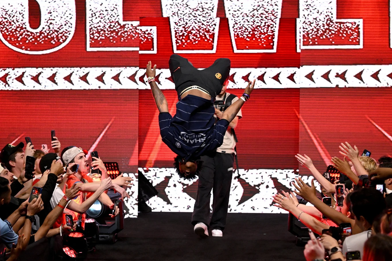 Mężczyzna wykonuje salto w tył na scenie podczas imprezy sportowej lub rozrywkowej, wokół tłum wiwatujących i fotografujących ludzi.