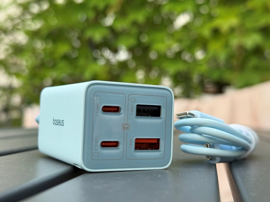 Ładowarka Baseus w jasnoniebieskim kolorze z czterema portami USB, w tym dwoma portami USB-C, jednym USB-A i jednym dodatkowym USB służąca do ładowania różnych urządzeń elektronicznych, leży na ciemnej ławce, obok niej widoczny jest kabel USB.
