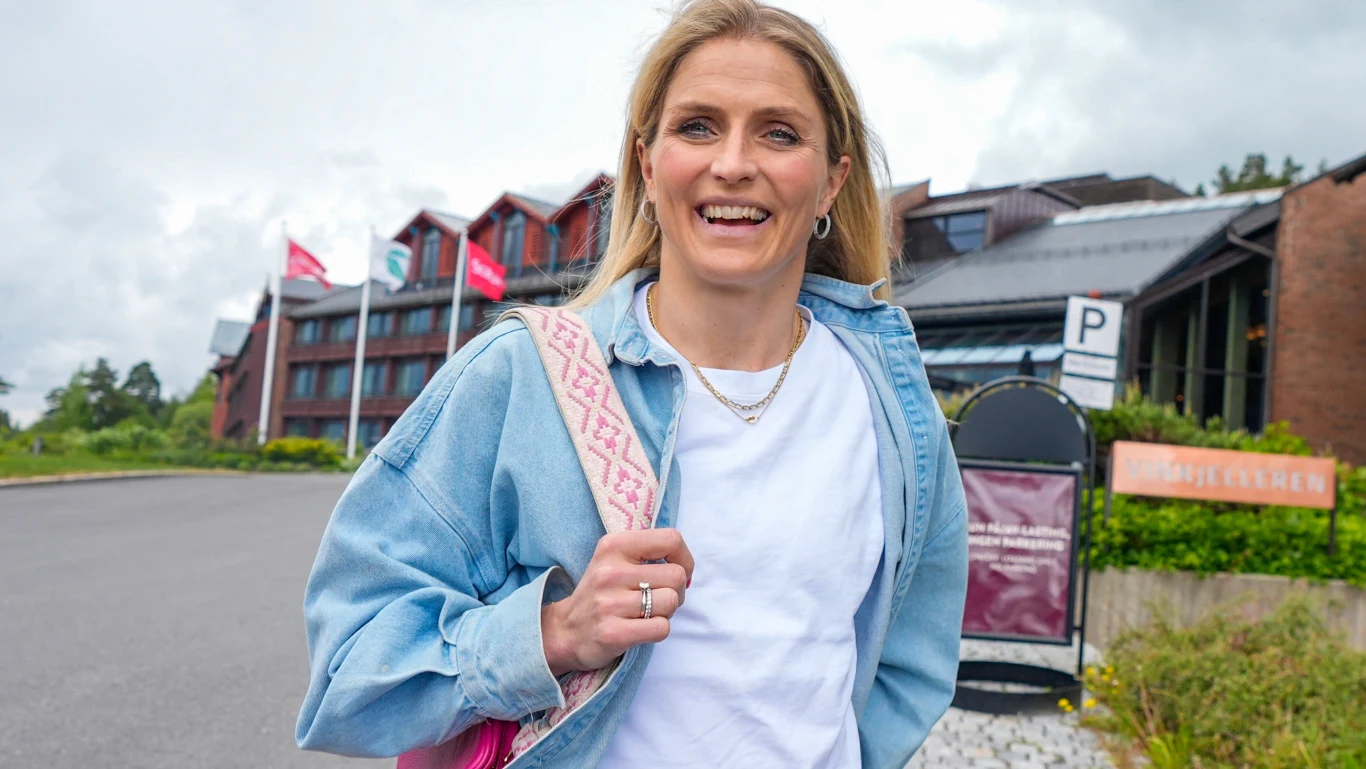 Kobieta z jasnymi włosami ubrana w białą koszulkę i niebieską kurtkę jeansową stoi na zewnątrz przed budynkiem z czerwonymi dachami, w tle widoczne flagi oraz wejście do obiektu.