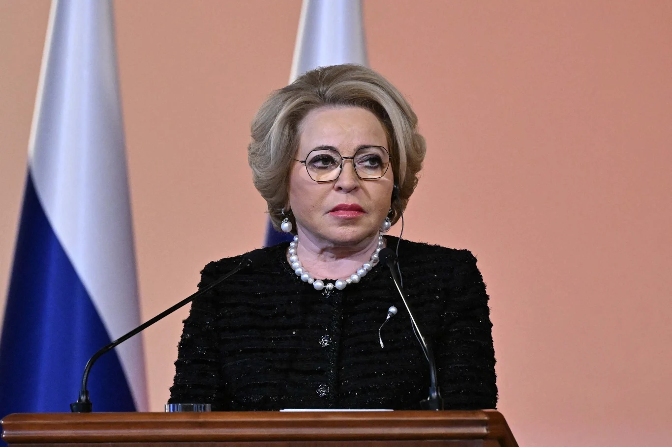 Walentina Matwijenko, mimo sankcji, przyleciała do Szwajcarii Starsza kobieta w okularach i eleganckiej czarnej marynarce stoi za mównicą, w tle widoczne dwie flagi biało-niebieskie. Ma jasne, starannie uczesane włosy oraz ozdobny naszyjnik z pereł.