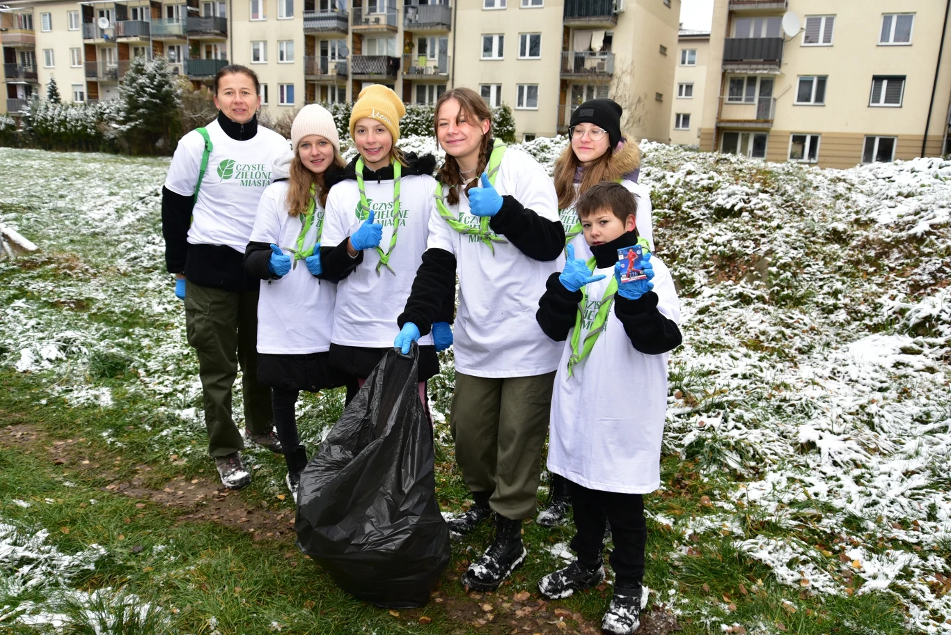 Grupa osób ubranych w białe koszulki z napisem Green Challenge, uśmiechnięte, trzymają worki na śmieci i rękawiczki, stoją na trawie częściowo pokrytej śniegiem na tle bloków mieszkalnych.