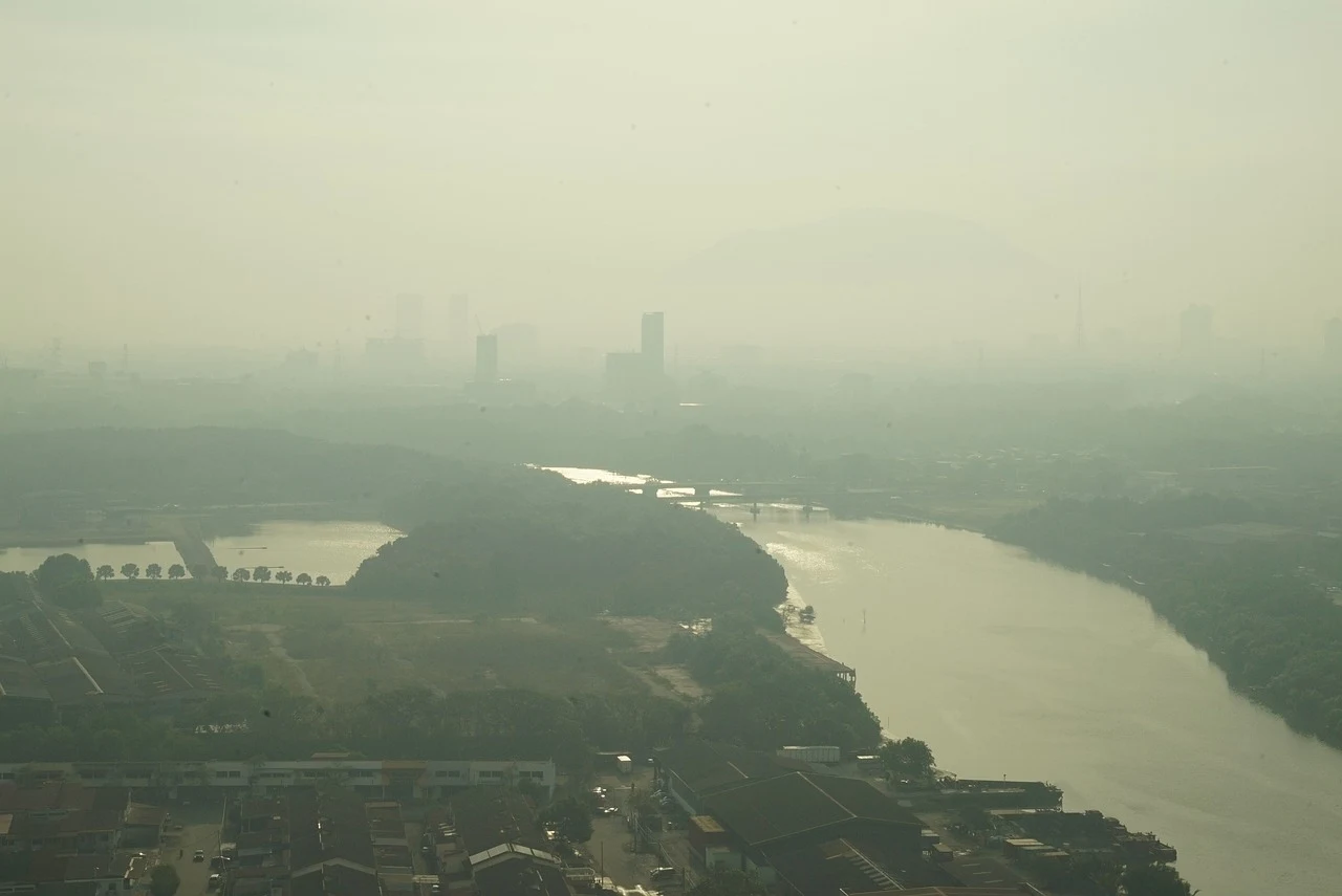 Panorama miasta zasnutego gęstym smogiem, widoczne zabudowania, rzeka wijąca się przez teren oraz ledwo widoczne w tle sylwetki wysokich budynków, niska widoczność spowodowana zanieczyszczeniem powietrza.