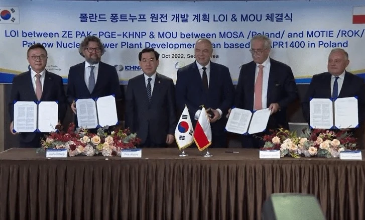 Grupa sześciu mężczyzn w garniturach stoi za biurkiem przykrytym brązowym obrusem, na którym ustawione są flagi Polski i Korei Południowej, kwiaty oraz dokumenty. Mężczyźni trzymają podpisane umowy w otwartych teczkach. W tle widoczny jest transparent ...