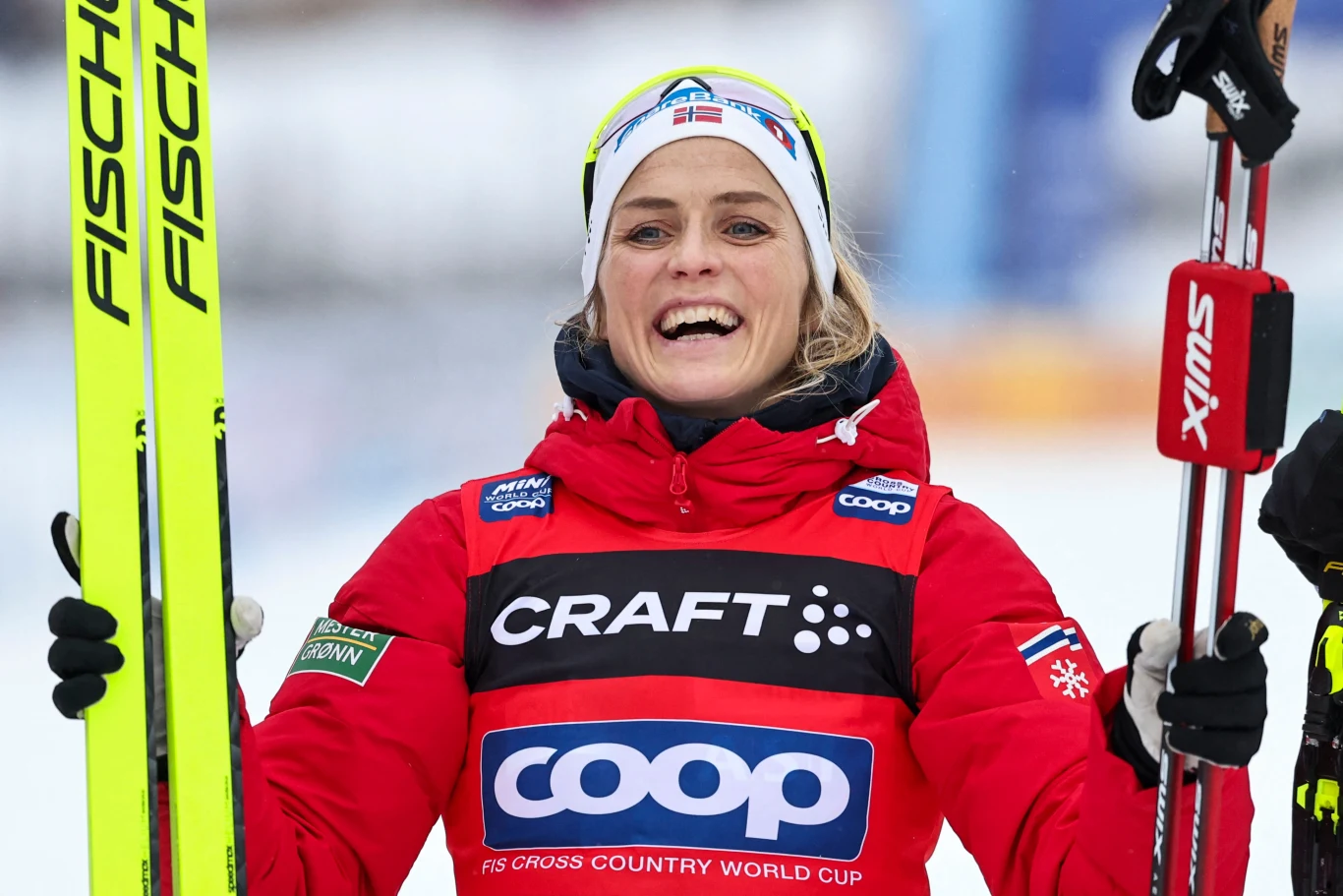 Zadowolona narciarka w czerwonej kurtce trzymająca narty i kijki, na głowie opaska z flagą kraju, w tle rozmyte zimowe otoczenie.