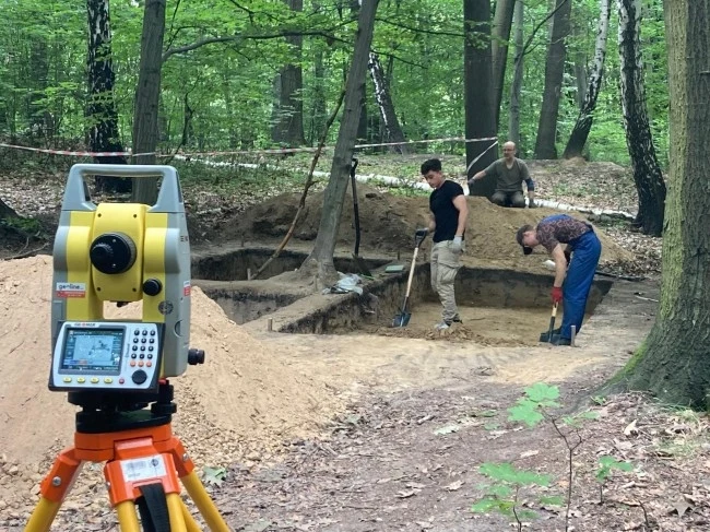 Prace archeologiczne prowadzone w lesie, trzech mężczyzn używa łopat podczas wykopalisk, w przednim planie urządzenie geodezyjne ustawione na statywie, wokół dużo drzew i zieleni.