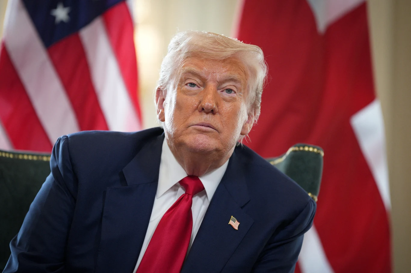 Mężczyzna w garniturze z czerwoną krawatą siedzi na tle flag Stanów Zjednoczonych oraz innego państwa, z poważnym wyrazem twarzy, patrząc lekko w bok.