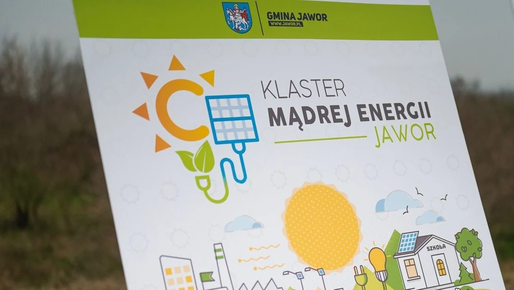 Tablica promująca inicjatywę Klaster Mądrej Energii Jawor z kolorową grafiką przedstawiającą elementy związane z energią odnawialną, takie jak słońce, panele słoneczne i domy.