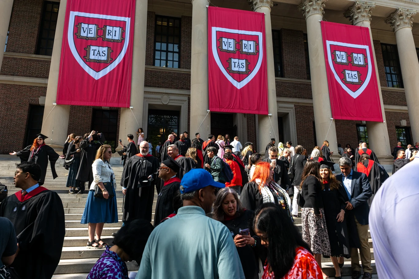 Grupa ludzi ubranych w togi i stroje akademickie zgromadzona na schodach przed budynkiem z trzema dużymi czerwonymi bannerami z napisem 'VERITAS' i herbem uniwersytetu.