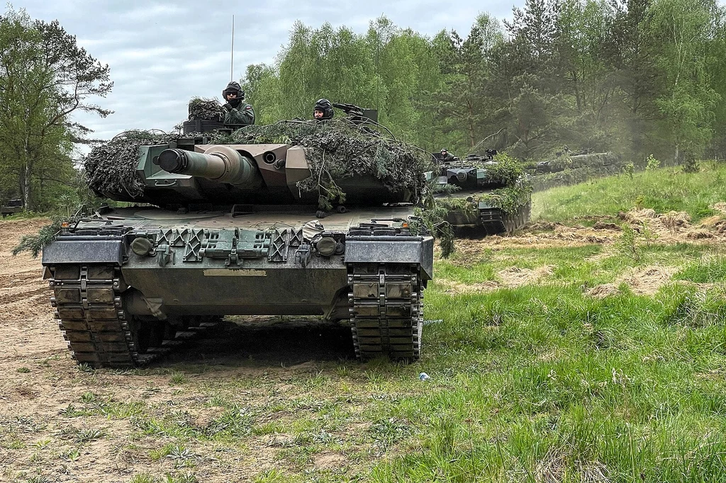 Polskie Leopardy 2PL Czołg pokryty siatkami maskującymi poruszający się po piaszczystym terenie, w tle drugi czołg częściowo zakryty przez drzewa, żołnierze w hełmach widoczni w wieżyczce, otoczenie stanowi zieleń lasu i trawa.