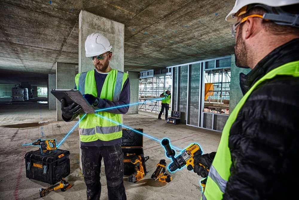 System DeWALT Tools Connect Dwóch pracowników budowlanych w kaskach i kamizelkach odblaskowych korzysta z tabletu oraz narzędzi elektrycznych, wykonując zadania w dużym, surowym wnętrzu budynku w trakcie budowy. W tle widoczna jest kolejna osoba wykonująca prace przy oknach.