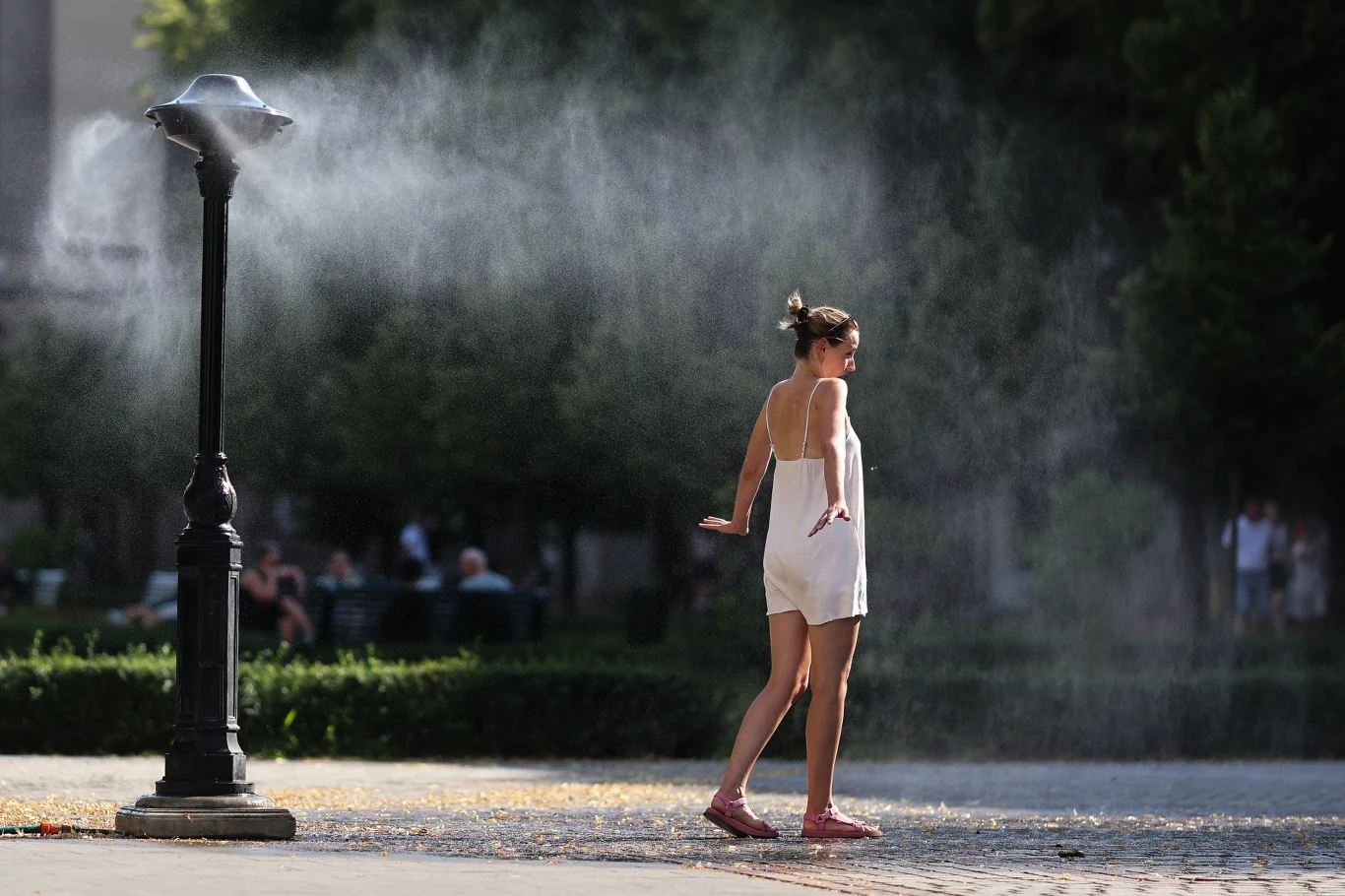 Upał w Krakowie (arch.) Kobieta w białej sukience ochładza się pod miejską mgiełką wodną emitowaną przez specjalną instalację w upalny dzień, otoczona zielenią i alejkami parku.