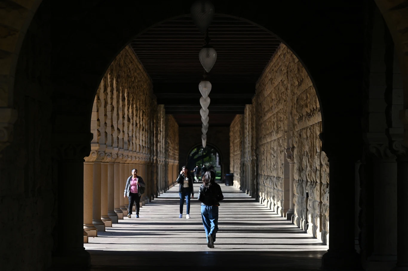 Kamienny korytarz z rzędami kolumn i dekoracyjnym sklepieniem łukowym, w którym kilka osób swobodnie spaceruje, światło dzienne tworzy wyraźne cienie na posadzce.